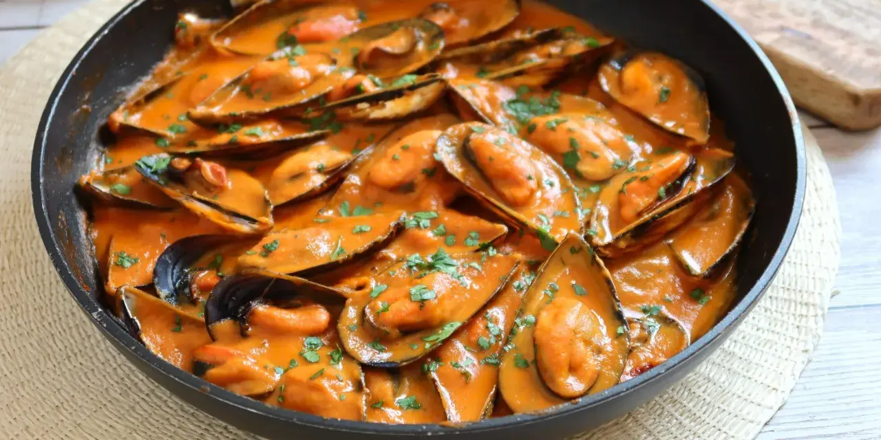 Mejillones con tomate en salsa cremosa, espolvoreados con perejil fresco, servidos en una sartén negra.