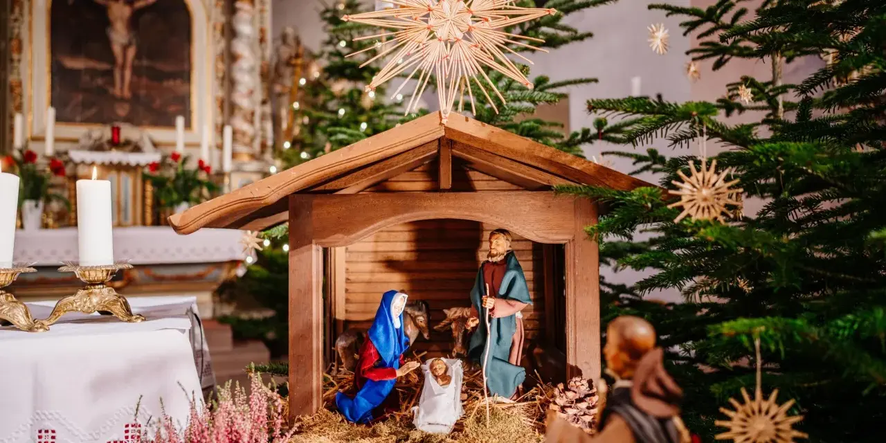 Weihnachtskrippe in einer Kirche, die die Geburt Jesu darstellt. Maria und Josef knien vor dem Kind, während im Hintergrund ein Weihnachtsbaum und Kerzen leuchten. So feiert man Weihnachten.