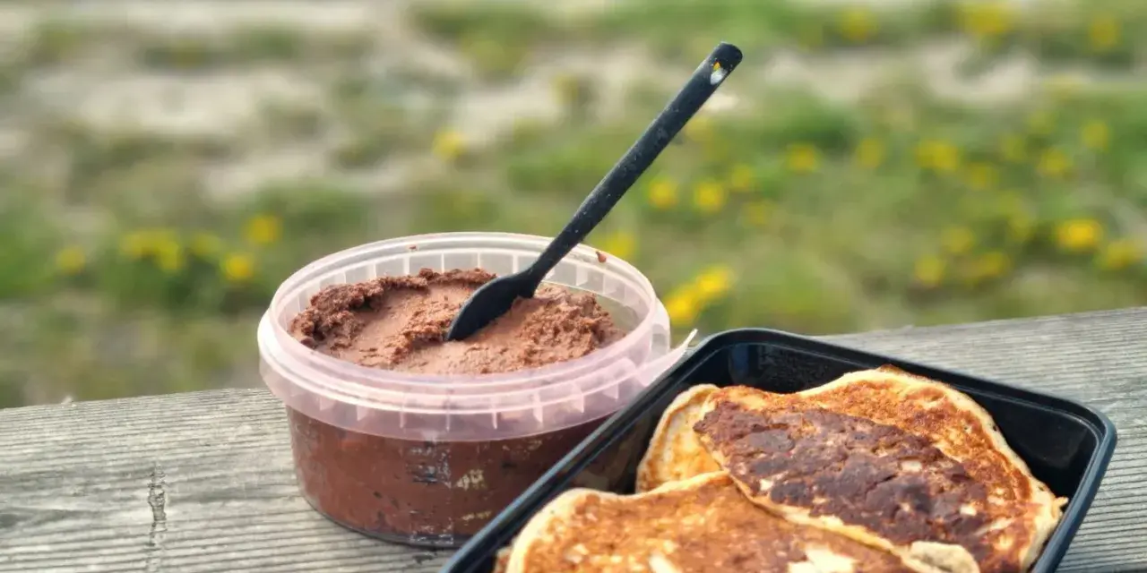 Färdiga proteinpannkakor i en svart behållare bredvid en burk med chokladpålägg, redo att avnjutas utomhus.