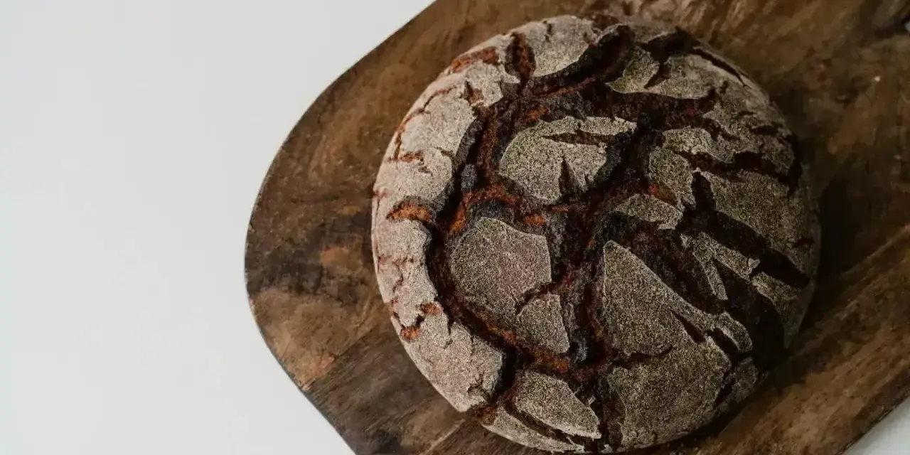 Chleb bez mąki, ciemny, okrągły bochenek z popękaną skórką, leży na drewnianej desce.