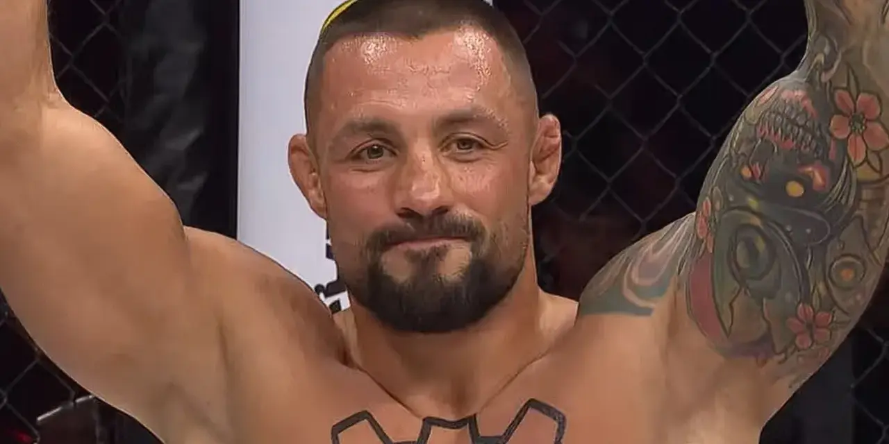 Mateusz Janur, wojownik MMA, z podniesionymi rękami i tatuażami na ramionach i klatce piersiowej, świętuje zwycięstwo.