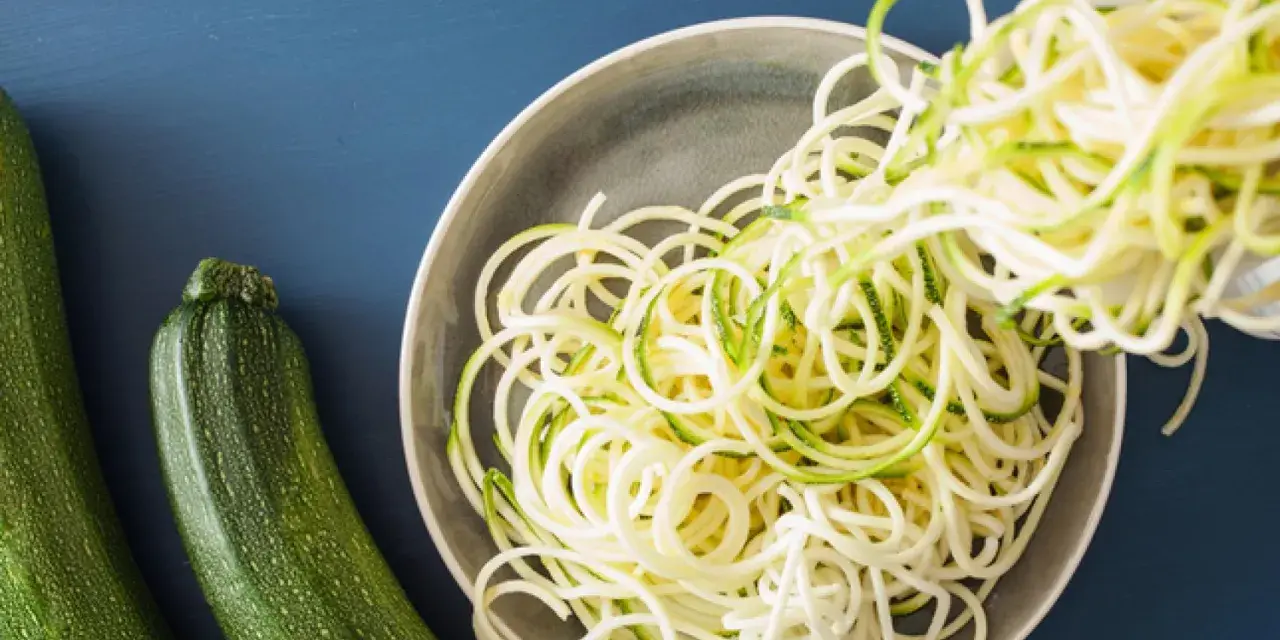 Espaguetis con calabacín recién hechos, listos para servir. Dos calabacines verdes reposan al lado de un plato lleno de fideos de calabacín.
