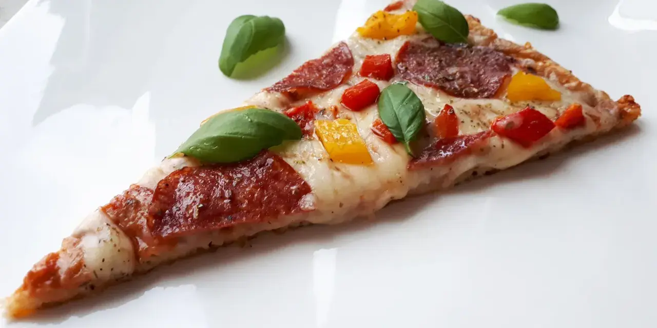 Kawałek pysznej pizzy keto z salami, papryką i bazylią na białym talerzu.