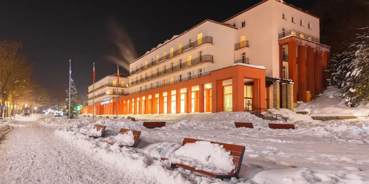 Zimowy wieczór w sanatorium godnym polecenia. Ośnieżone ławki i oświetlony budynek zapraszają do odpoczynku.