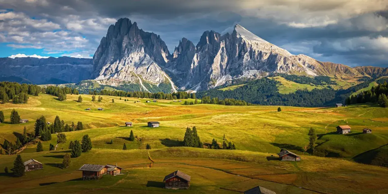 Un paesaggio alpino con un sasso lungo e imponente che domina un sasso piatto e verdeggiante, punteggiato da casette di legno.