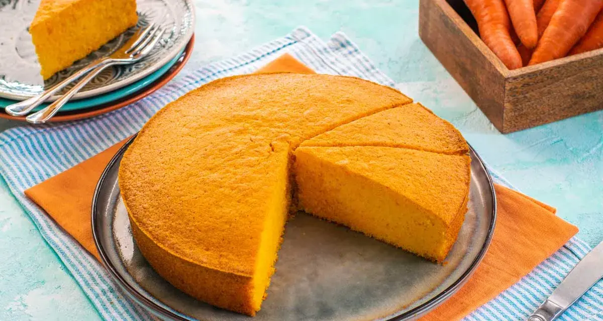 Una fetta di torta di carote sofficissima, appena tagliata da una torta intera su un piatto, con carote fresche in un cesto sullo sfondo.
