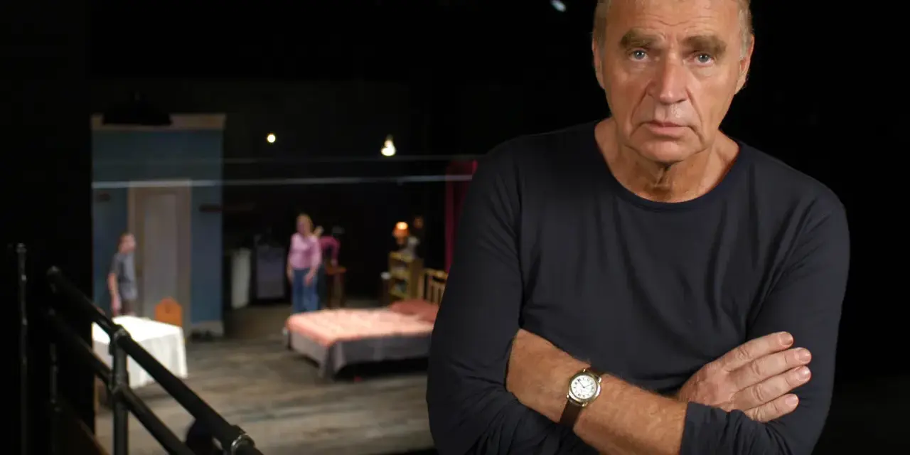 Dawid Głowacki, reżyser teatralny, z rękami skrzyżowanymi na piersi, na tle sceny z łóżkiem i ludźmi.