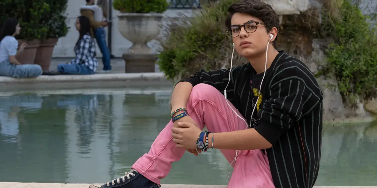 Il ragazzo dai pantaloni rosa, con occhiali e auricolari, ascolta musica seduto vicino a una fontana, mentre altre persone chiacchierano sullo sfondo.
