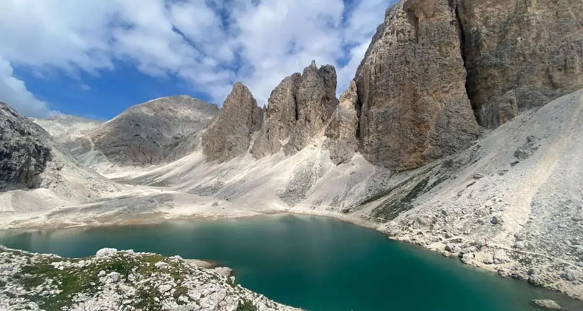 Il **lago di Antermoia**, incastonato tra imponenti vette rocciose, riflette il cielo azzurro e le nuvole bianche. Un gioiello alpino.
