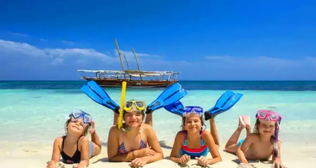 Cztery uśmiechnięte dzieci w maskach do snorkelingu i płetwach leżą na plaży. W tle turkusowa woda i łódź. Idealny dzień, kiedy na Zanzibar!