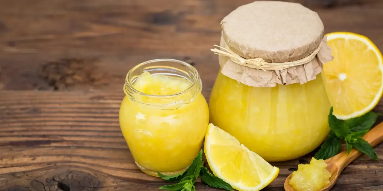 Due vasetti di marmellata di limoni, limoni freschi e foglie di menta su un tavolo di legno.