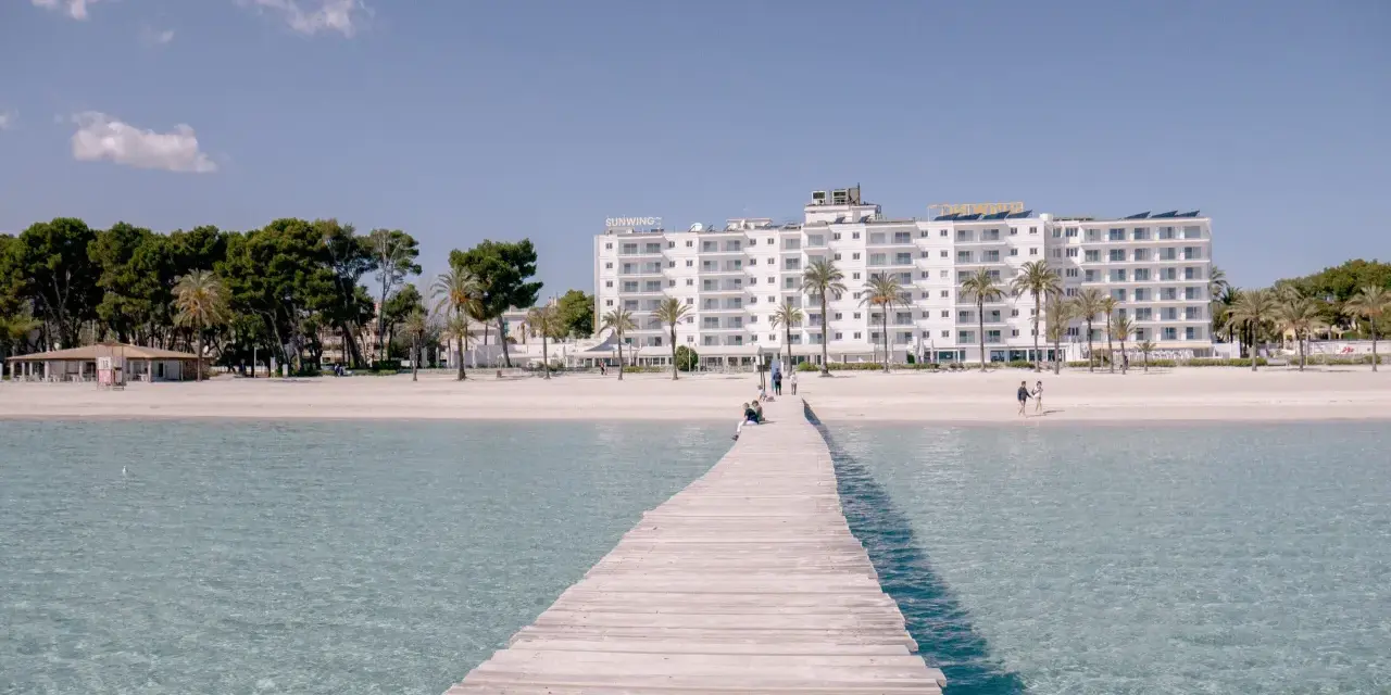 Drewniany pomost prowadzi do piaszczystej plaży Playa de Muro z białym hotelem i palmami w tle.