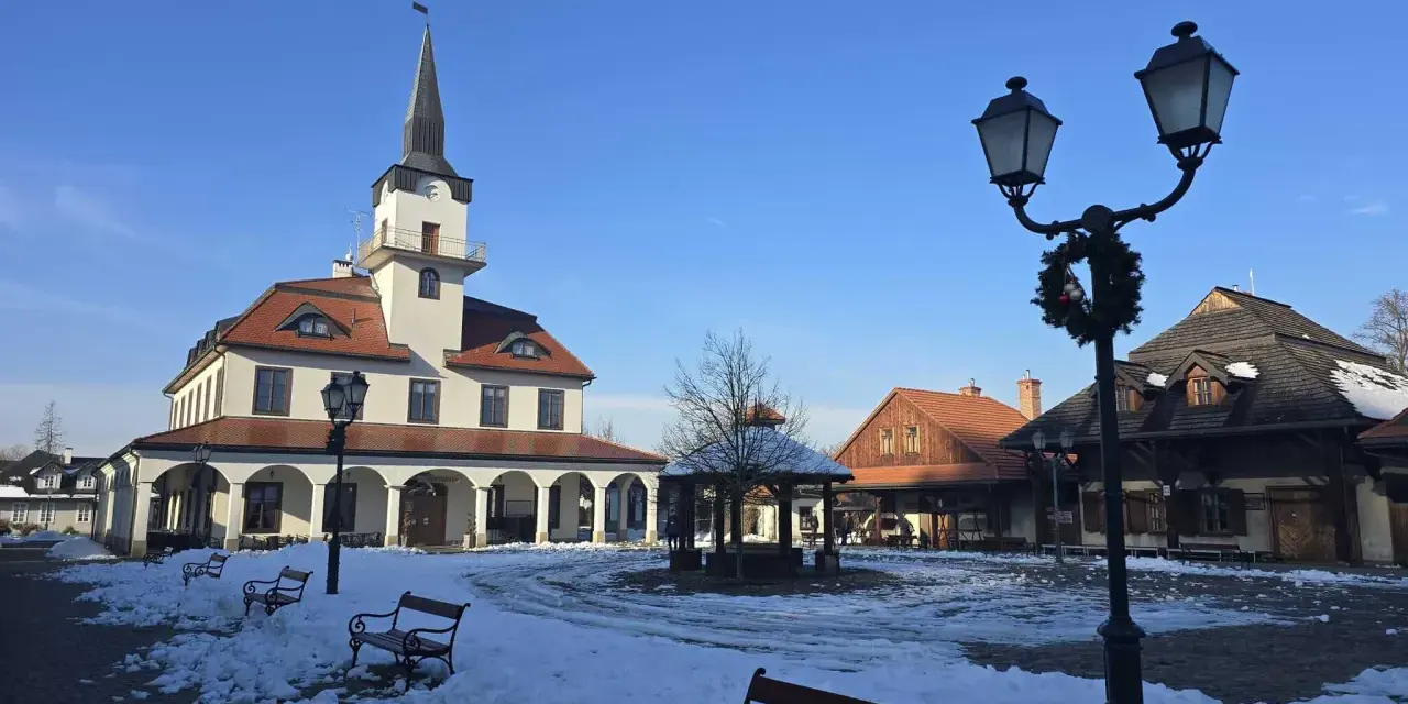 Zimowe miasteczko galicyjskie z ratuszem i karczmą, plac pokryty śniegiem, ławki i latarnie ozdobione świąteczną girlandą.