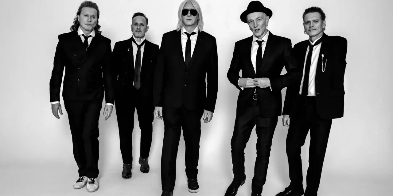 Fünf Männer in schwarzen Anzügen und Krawatten, die wie der titel von Def Leppard aussehen, posieren für ein Schwarz-Weiß-Foto.