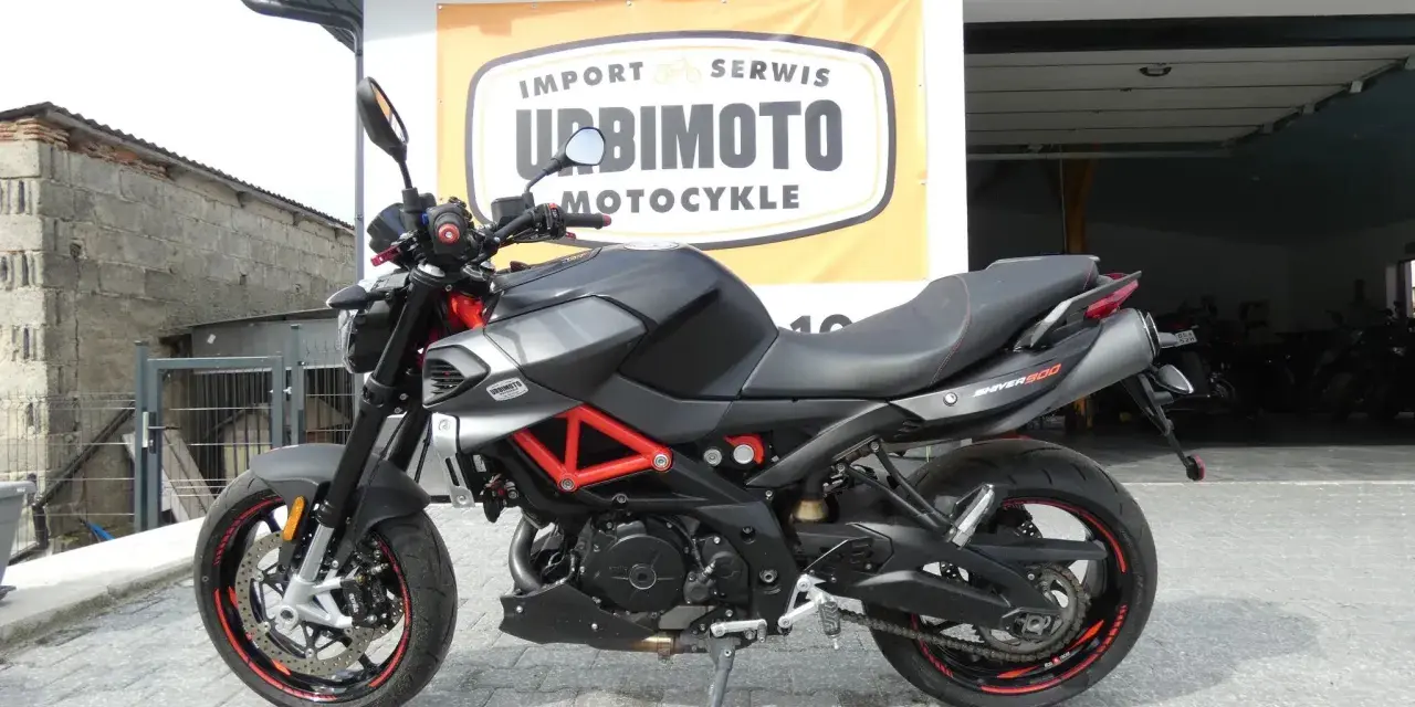 Czarny motocykl Aprilia Shiver 900 z czerwonym stelażem, zaparkowany przed salonem motocyklowym.