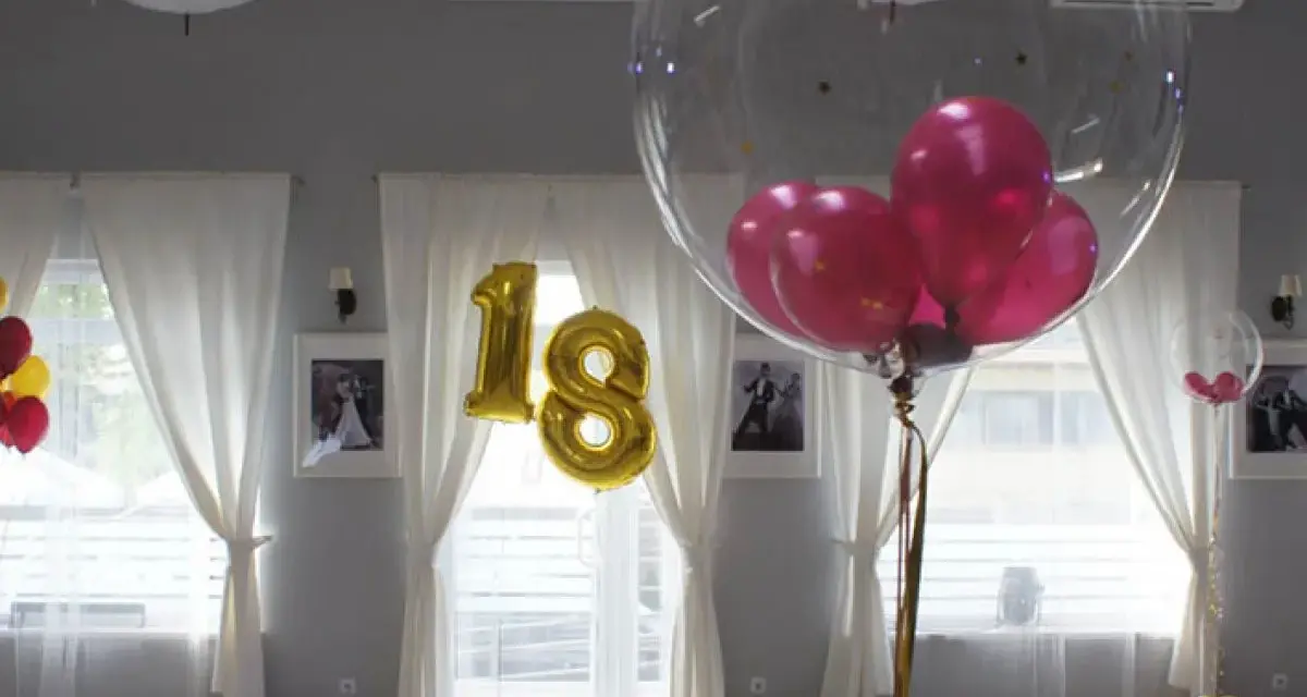 Złote balony z cyfrą 18 i przezroczyste balony z różowymi w środku ozdabiają salę. Balony na stołach tworzą radosną atmosferę.