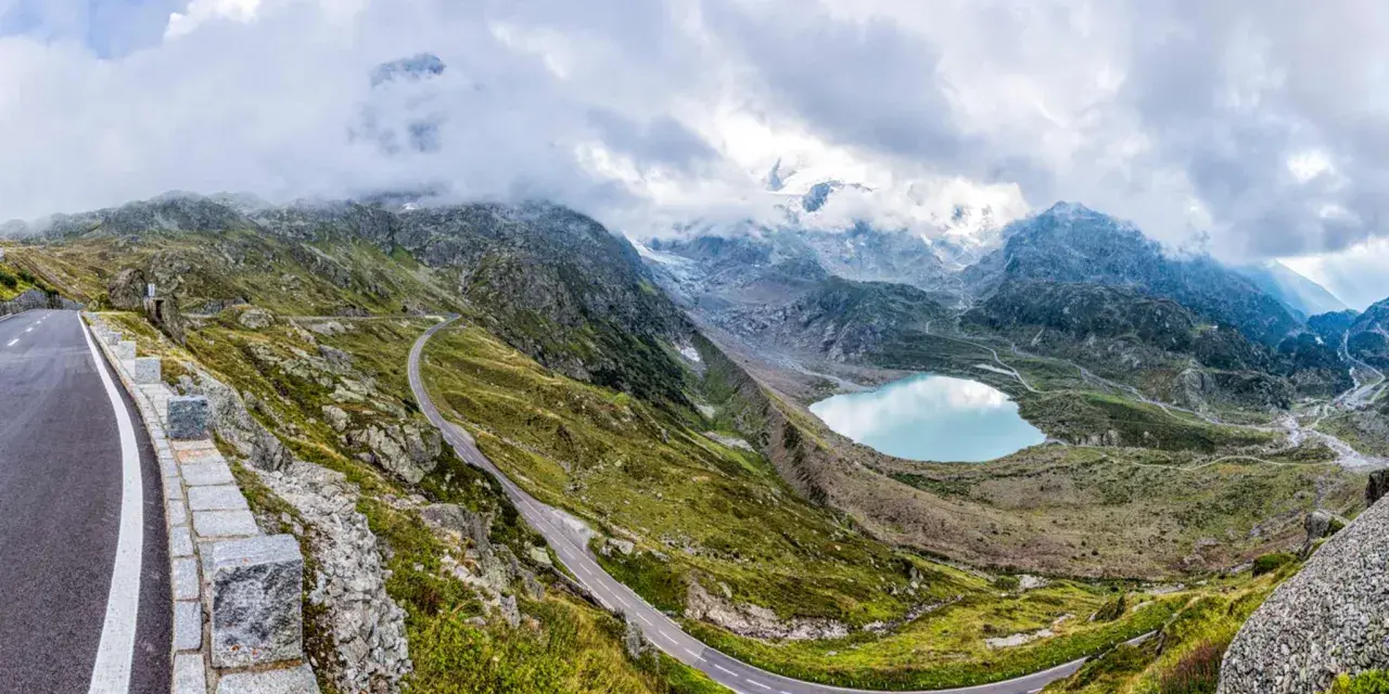 Strada tortuosa sul Passo San Lugano, con lago alpino azzurro e montagne nebbiose sullo sfondo.