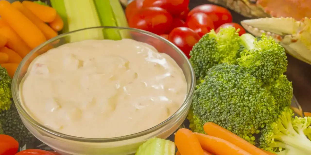 Un bol de sauce crudités crème fraîche entouré de carottes, céleri, tomates cerises et brocolis. Un crabe cuit est visible en arrière-plan.