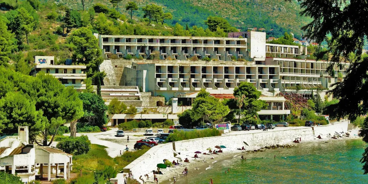 Opuszczone hotele Chorwacja, zniszczone budynki hotelowe na wzgórzu, plaża z ludźmi i morze.