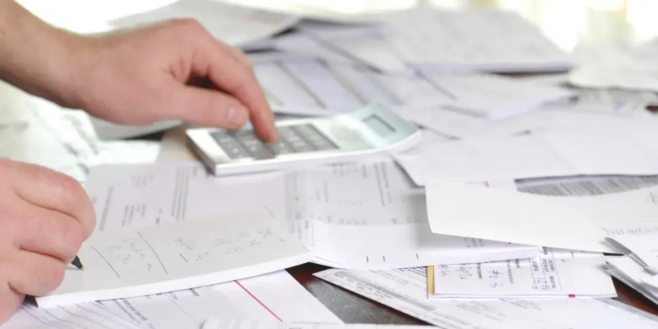 Mano calculando y anotando en libreta, rodeado de papeles y facturas con retención.