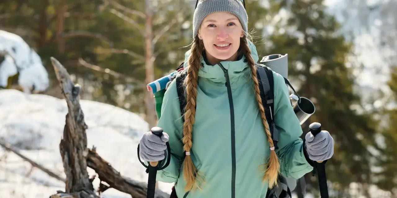 Donna con trecce e zaino, pronta per attività invernali, sorride in un paesaggio innevato con bastoncini da trekking.