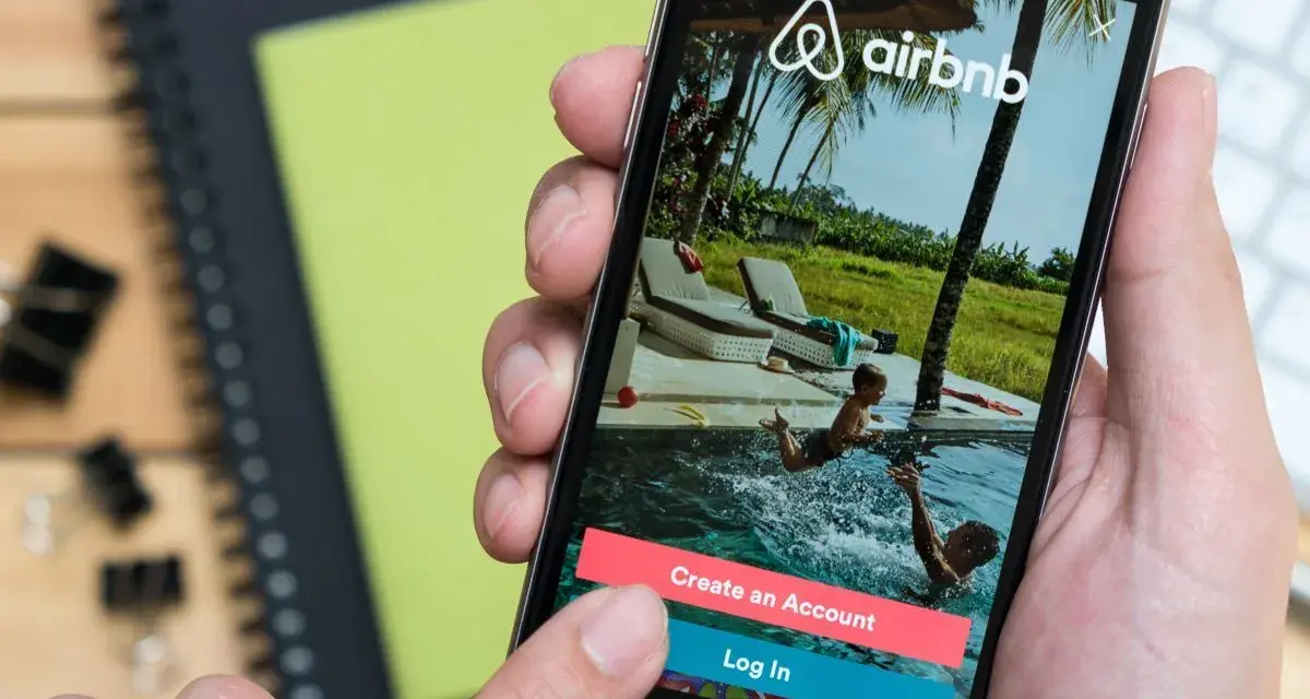 Dłoń trzyma smartfon z aplikacją Airbnb. Czy Airbnb jest bezpieczne? Na ekranie widać basen i dzieci, przyciski "Create an Account" i "Log In".