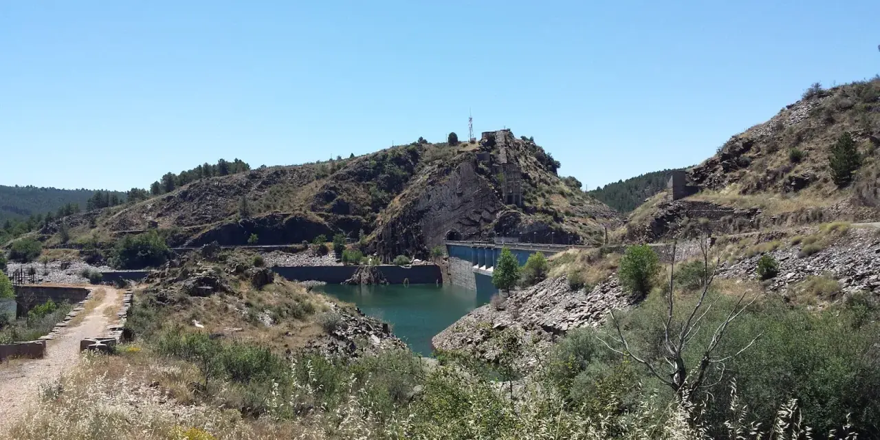 El embalse del Vado, con su presa de hormigón y aguas turquesas, se abre paso entre colinas rocosas y vegetación seca bajo un cielo azul.