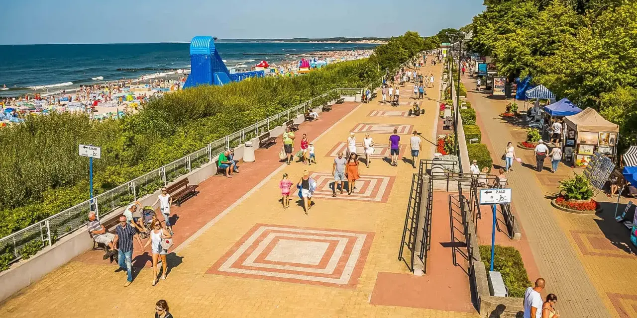 Tętniąca życiem ustka promenada w słoneczny dzień. Ludzie spacerują, odpoczywają na ławkach, a w tle widać plażę pełną turystów i morze.