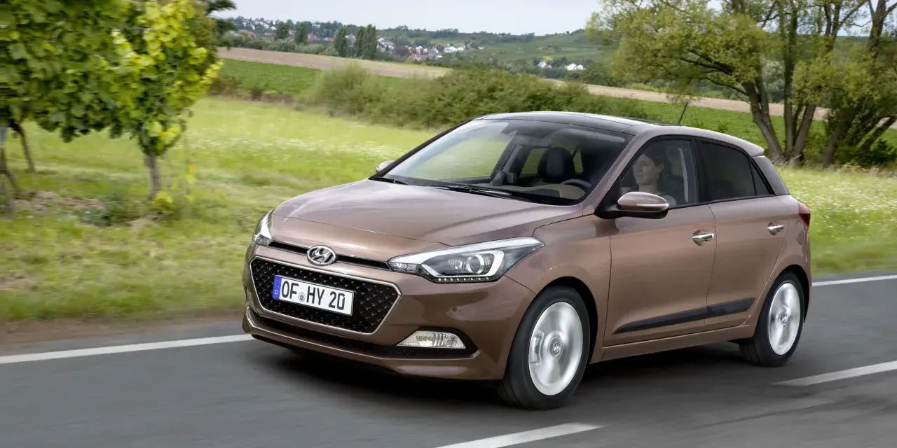 Brązowy Hyundai i20 na drodze wśród zieleni. Kierowca uśmiecha się, co może sugerować pozytywne hyundai i20 opinie.