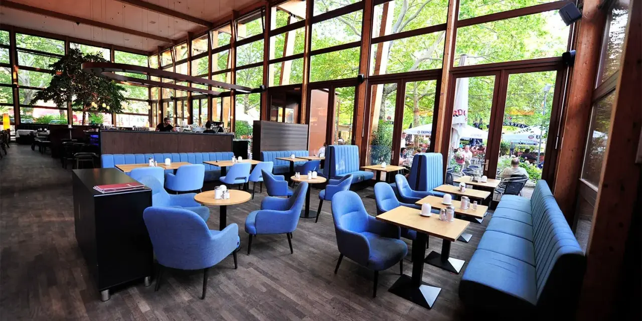 Gemütliches **Cafe Wilmersdorf** mit blauen Sesseln und Bänken, Holzboden und großen Fenstern, die auf eine grüne Terrasse blicken.