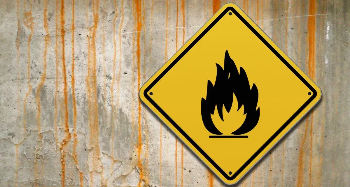 Cartello giallo con fiamma nera su muro di cemento con macchie arrugginite. Attenzione: lana di roccia è infiammabile.