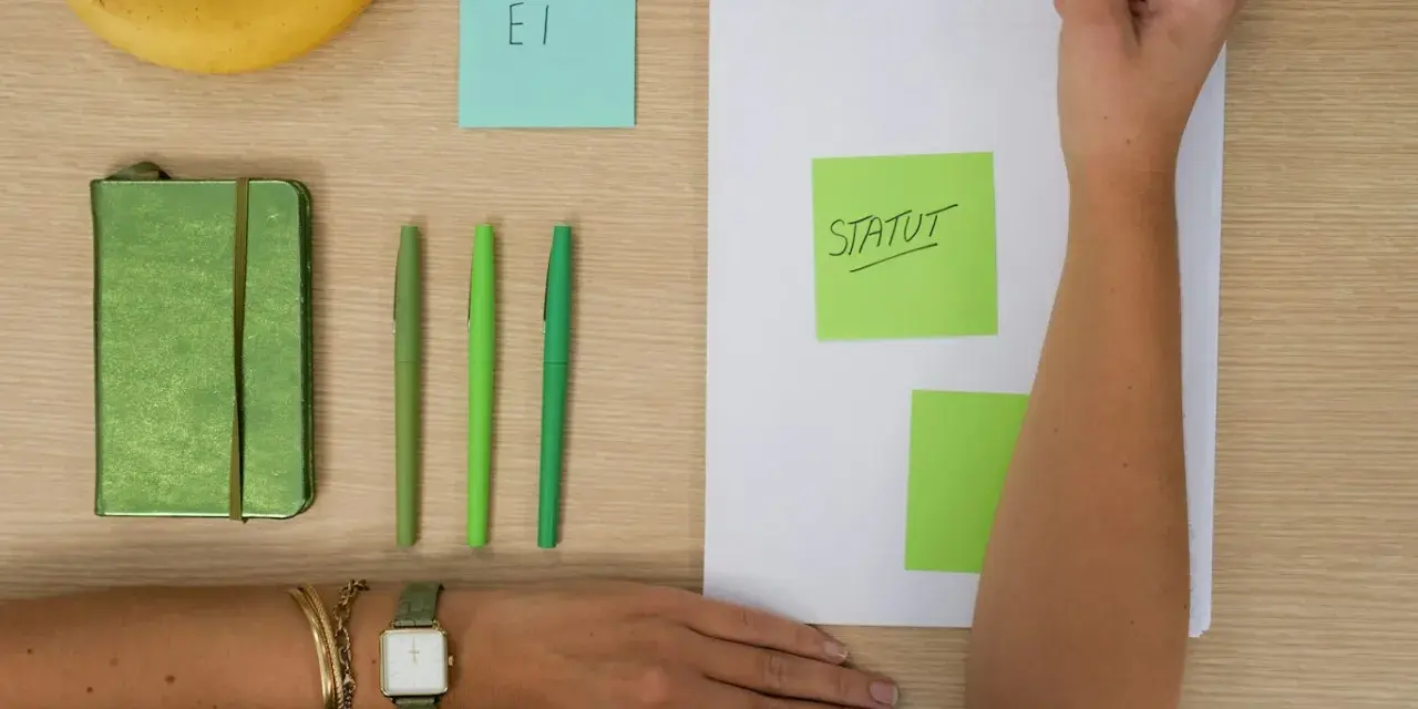 Une main écrit "STATUT" sur un post-it vert, à côté d'un carnet, de stylos et d'une banane. L'acronyme "EI" sur un post-it bleu suggère une **entreprise individuelle**.