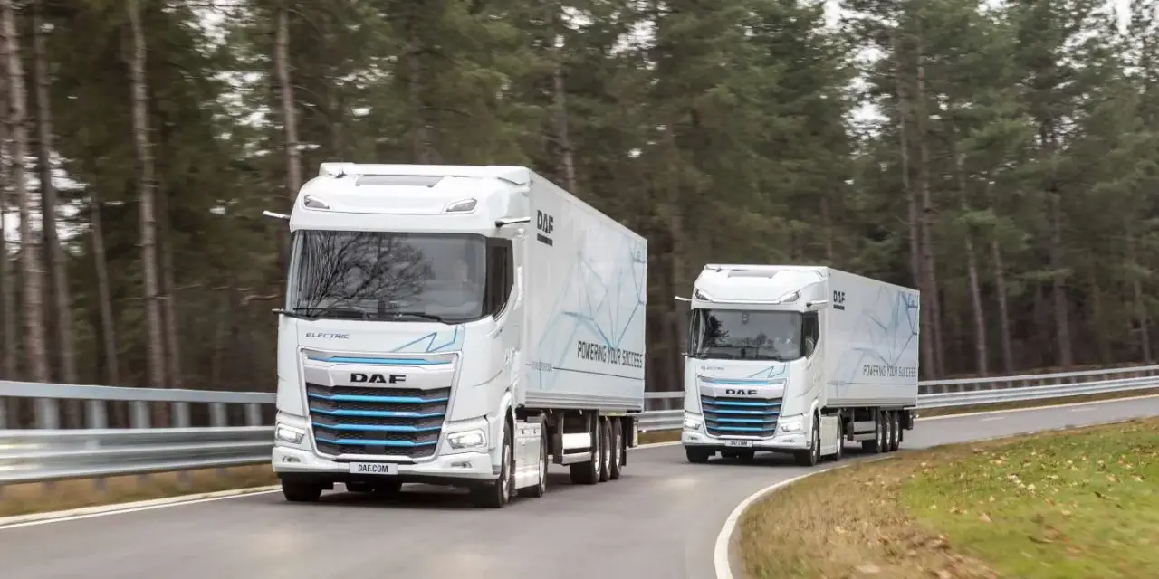 Dwa białe elektryczne DAF XG Plus z naczepami jadące po krętej drodze wśród drzew.