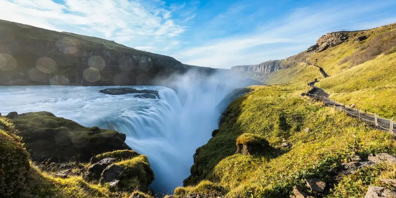 Potężny wodospad Gullfoss w Islandii, z mgiełką unoszącą się nad kaskadami. Ścieżka turystyczna wije się po zielonym zboczu.