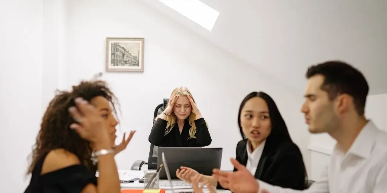 Un gruppo di colleghi di lavoro discute animatamente. Una donna bionda sembra stressata, tenendosi la testa tra le mani.