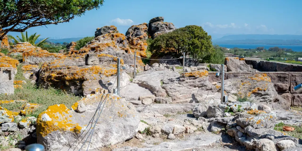 Ancient Italian ruins with weathered stones covered in orange lichen, a pathway winding through the site, and a distant blue sea under a clear sky.