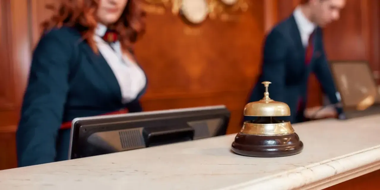 Recepcjonistka i recepcjonista za ladą hotelową. Dzwonek do obsługi gości, który pomoże dowiedzieć się, ile kosztuje doba hotelowa.