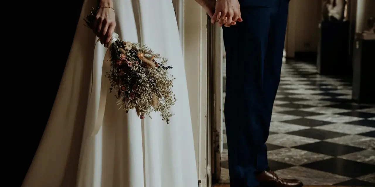 Para młoda trzyma się za ręce, ona z bukietem suszonych kwiatów. Wesele motywy w eleganckim wnętrzu.