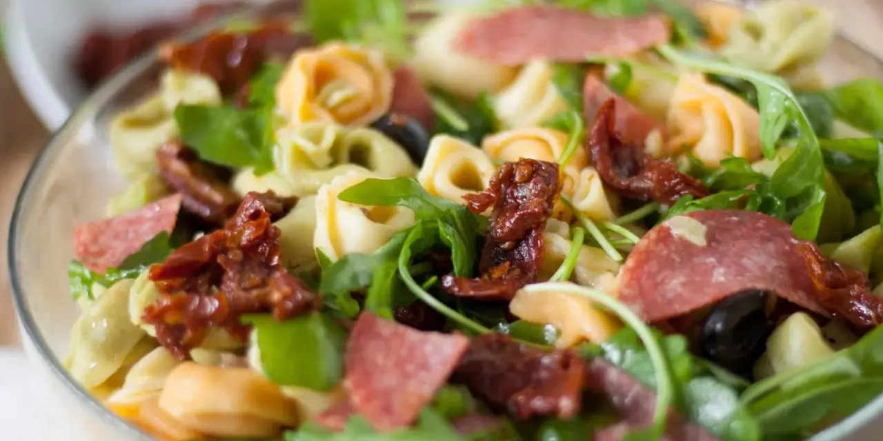 Sałatka z tortellini, suszonymi pomidorami, rukolą i salami w szklanej misce.