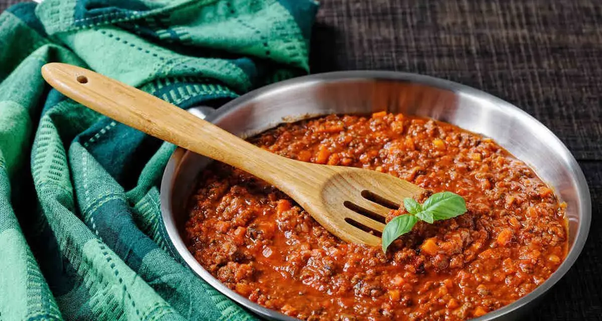 Un saporito ragù di cinghiale in padella, pronto per condire la pasta. Un cucchiaio di legno e una fogliolina di basilico completano la scena.