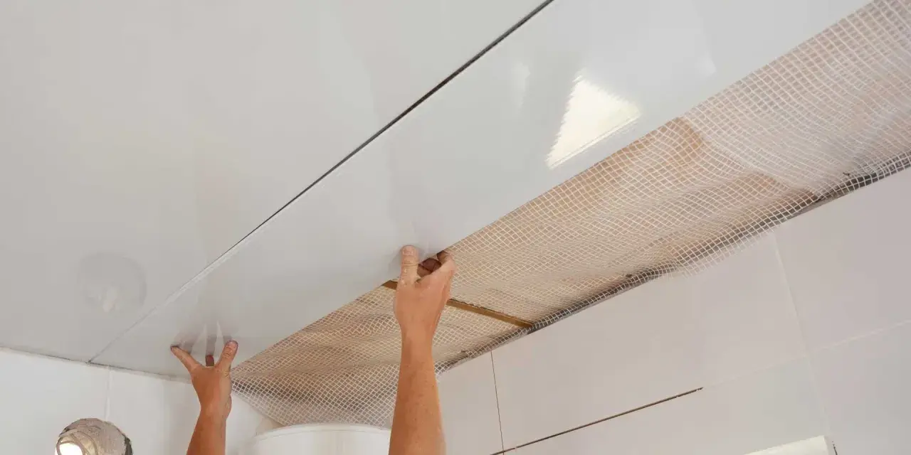 Pose de lambris PVC au plafond avec clips. Les mains ajustent une lame blanche brillante sur une structure, révélant un treillis blanc en dessous.