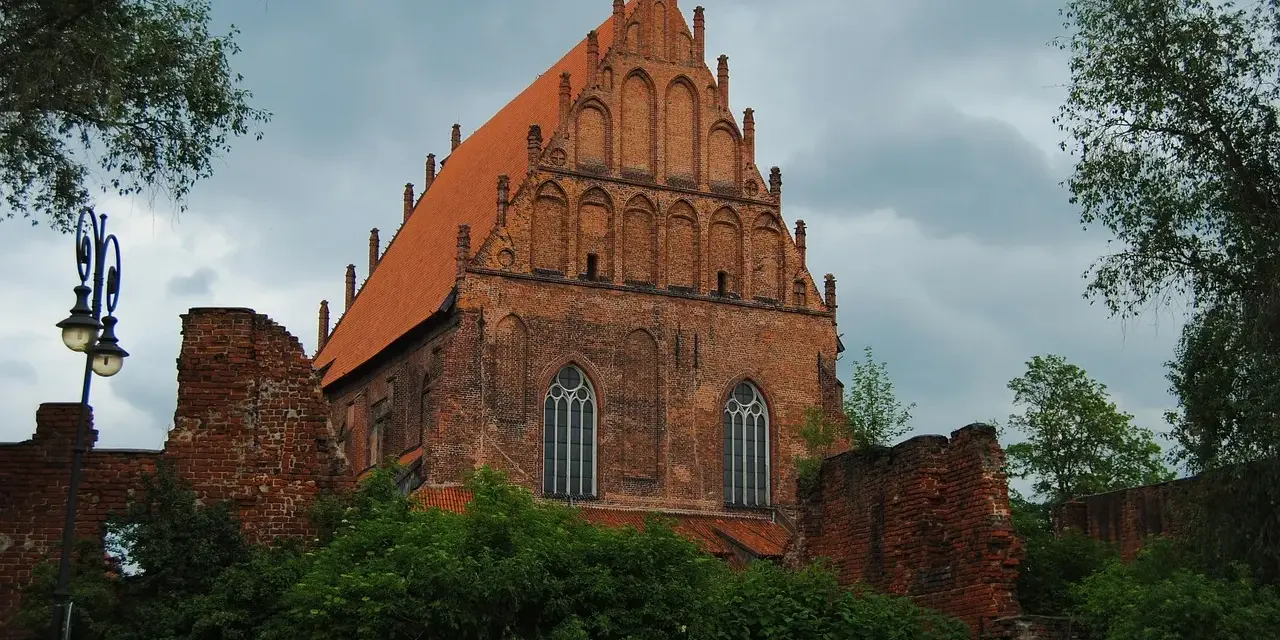 Gotycka ceglana budowla z pomarańczowym dachem, otoczona zielenią i fragmentami murów. Widok na Elbląg, województwo warmińsko-mazurskie.