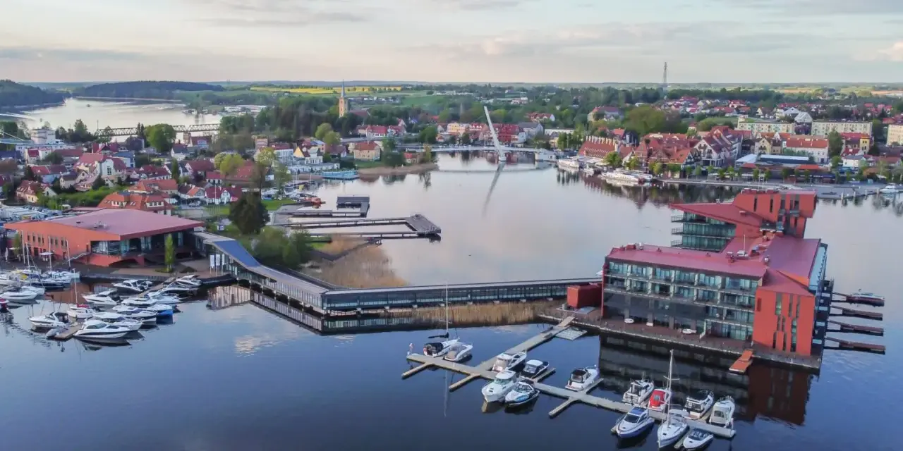 Nowoczesna architektura nad jeziorem, z łodziami przycumowanymi do pomostów. To jedno z **ciekawych miejsc na Mazurach**, idealne na wypoczynek.