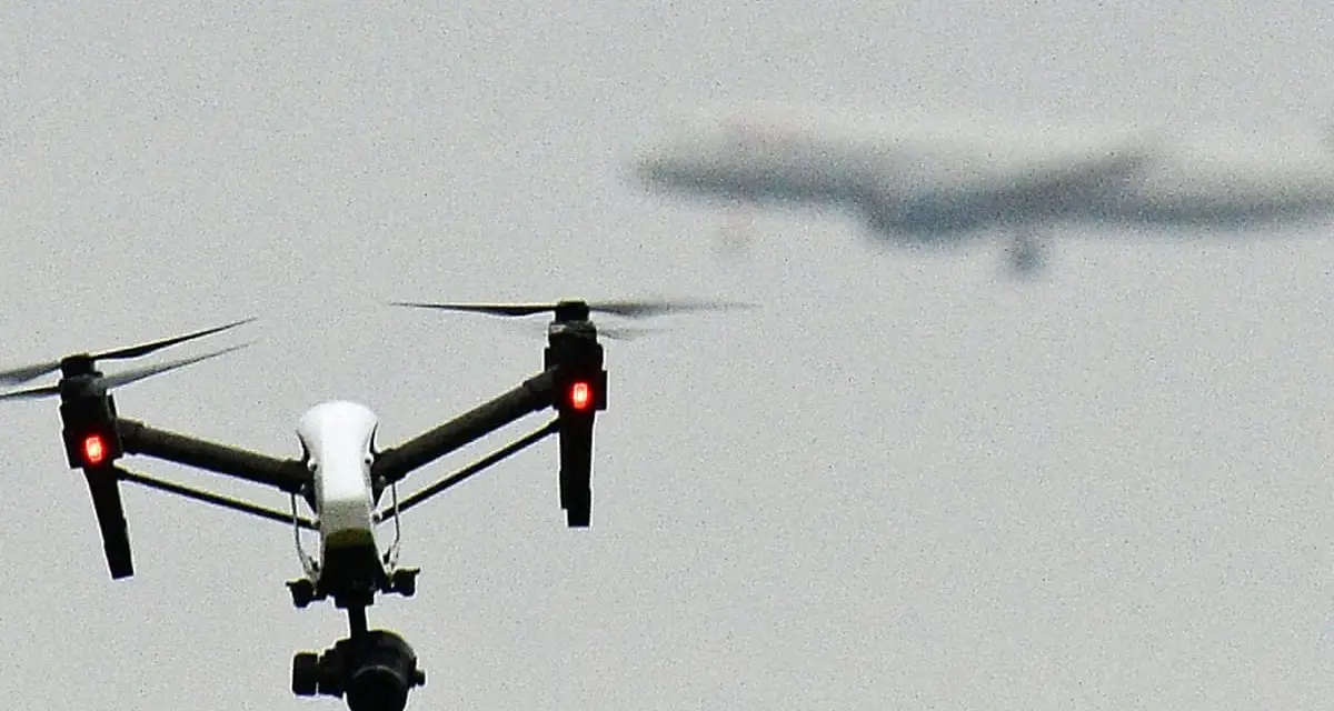 A white drone with red lights hovers in the sky, possibly on a security mission, as a blurred airplane flies in the distance.