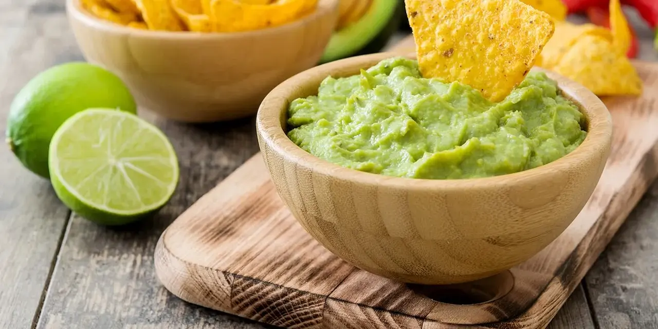 Pyszne guacamole w drewnianej misce, idealne do maczania nachos. Obok limonki i miska pełna chipsów. Z czym jeść guacamole? Z nachos!