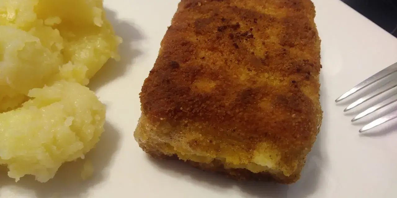 Chrupiąca panierowana kostka rybna z purée ziemniaczanym na białym talerzu.