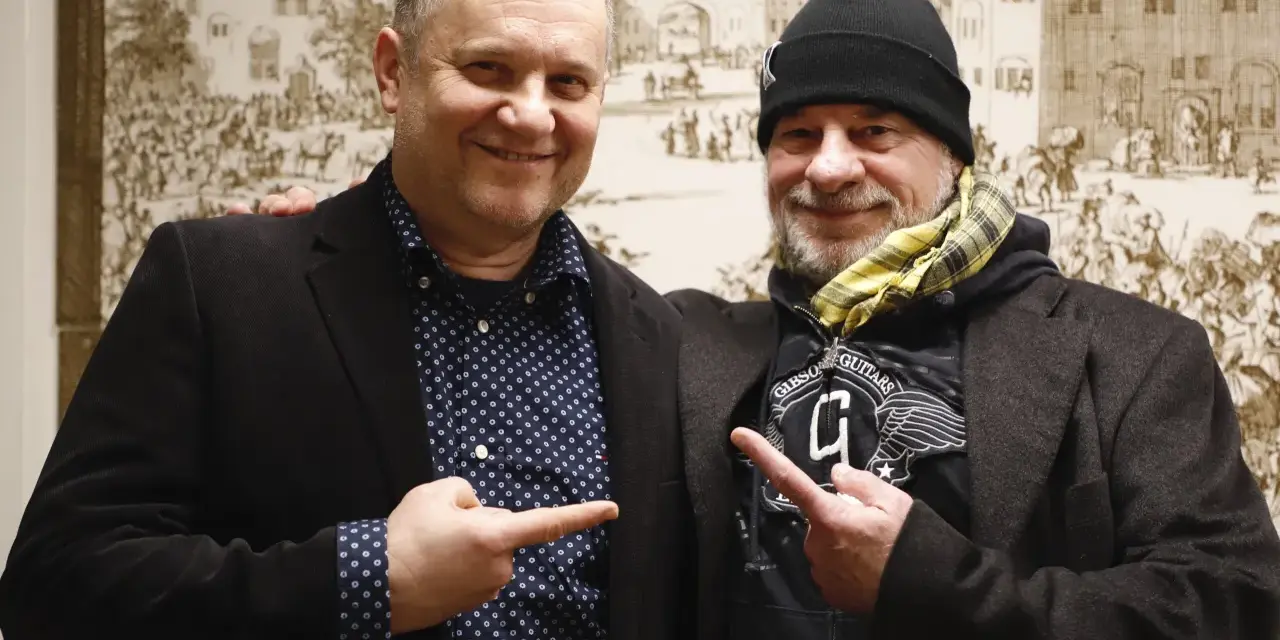 Deux hommes souriants, l'un avec une chemise à pois, l'autre avec un bonnet, se montrent du doigt. Un chanteur de confiance.