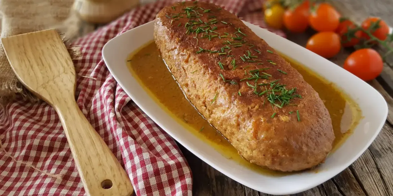 Polpettone in padella dorato, guarnito con erba cipollina, servito su un piatto bianco con salsa. Accanto, una spatola di legno e pomodorini.
