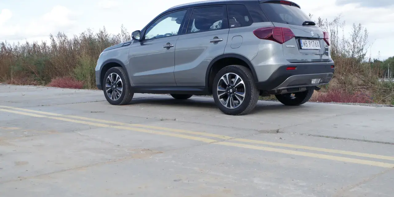 Szary Suzuki Vitara Hybrid z czarnym dachem stoi na betonowej nawierzchni z żółtymi pasami.
