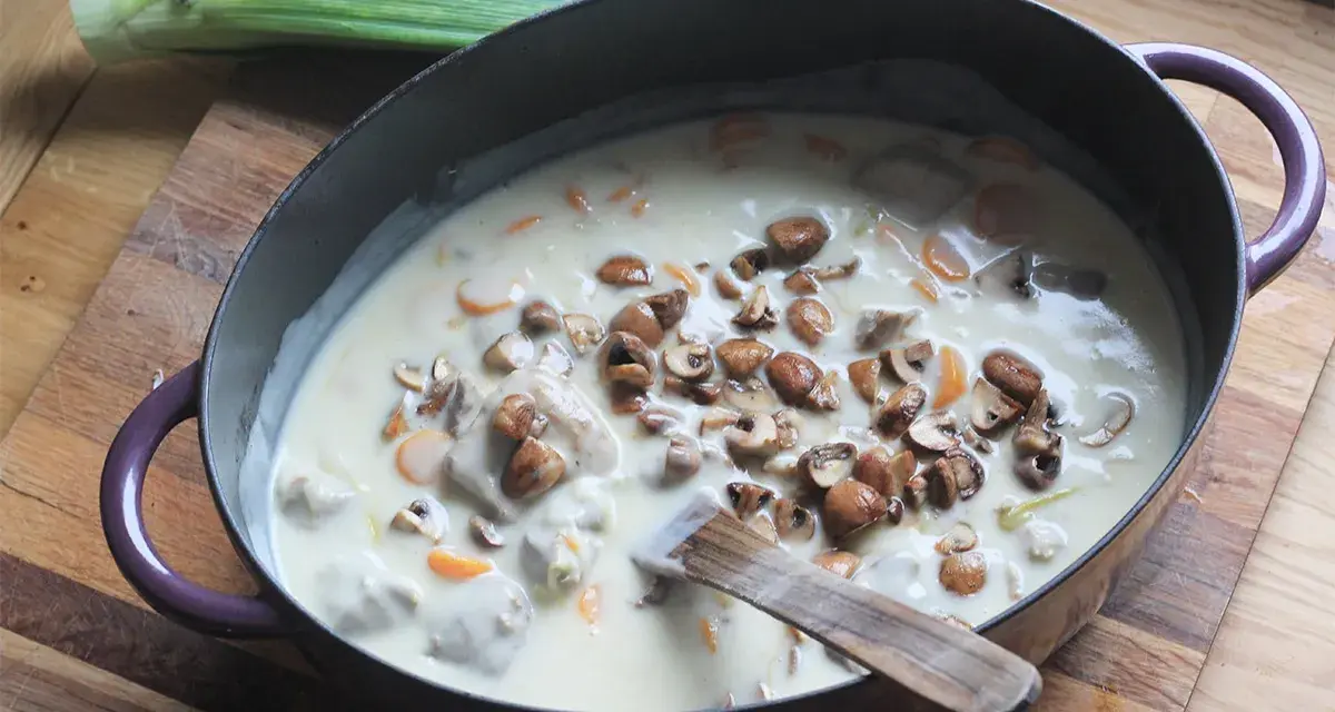 Un plat de blanquette de veau facile mijote dans une cocotte, garni de champignons et de carottes. Un poireau est posé à côté.